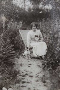 Photo of Hilda Burkitt sitting on a deck chair in a garden. She is wearing a white dress and has dark curly hair. She's stroking a small dog that's standing beside her.